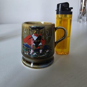 May include: A ceramic mug with a handle, featuring a colorful illustration of a figure carrying a basket. The mug has a dark green and brown color scheme. A yellow lighter is next to the mug.