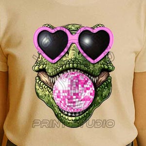 Op de afbeelding: Wit T-shirt met een groene dinosaurus die een roze hartvormige zonnebril draagt en een roze bel blaast met een discobal erin. De tekst "PRINT STUDIO" staat onderaan het shirt gedrukt.