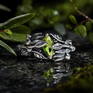 May include: A silver ring featuring a marquise-cut lime green gemstone set in the center. The band is designed with detailed metal leaves and small berries wrapping around the stone, creating a textured, organic appearance in the metalwork.