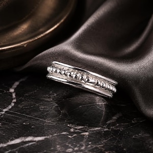May include: A silver ring featuring a central band with a textured, beaded, or rope-like pattern flanked by two smooth, polished silver rails on the top and bottom edges, creating a layered and dimensional appearance with a reflective metallic finish.