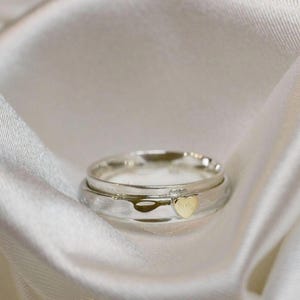 Puede incluir: Un anillo de plata con un detalle de corazón dorado. El anillo se presenta sobre un fondo de satén blanco, creando una sensación suave y romántica. El diseño del anillo es simple pero elegante, adecuado para el uso diario o ocasiones especiales.