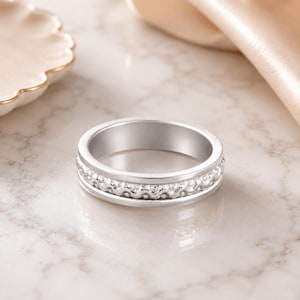 May include: A silver ring with a decorative band featuring a repeating pattern of small, raised elements. The ring is set against a soft, neutral background, highlighting its polished finish and intricate design. The ring's band is approximately 0.64 cm wide.