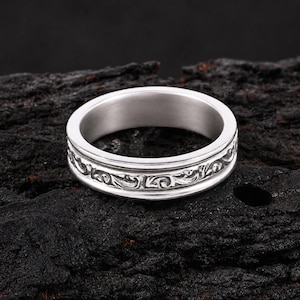 Puede incluir: Un anillo plateado con un intrincado diseño floral alrededor de la banda. El anillo, hecho de un material metálico brillante, se encuentra sobre una superficie oscura y texturizada. El diseño es detallado y elegante.