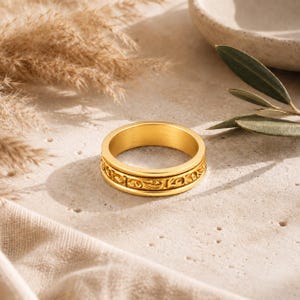 May include: A gold-colored ring with an engraved floral pattern around the band. The ring is set against a neutral background with dried plants and a ceramic bowl.