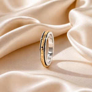 May include: A two-tone ring featuring a central gold-coloured band with a hammered texture, flanked by two thin black grooves and smooth silver-coloured edges. The ring stands upright, showcasing its layered metallic design and polished finish.
