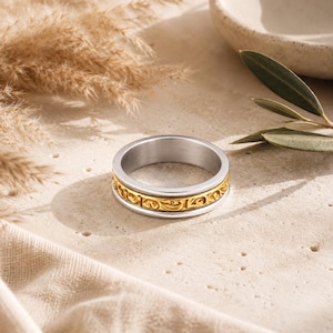 May include: A silver ring with a gold band featuring an ornate, repeating pattern. The ring is displayed on a neutral surface, with dried grasses and a ceramic bowl in the background. The ring's design is detailed and eye-catching.