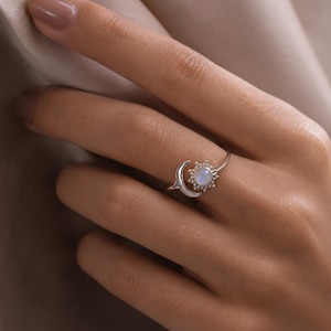 May include: A silver ring featuring a celestial design. The ring has a crescent moon on one side and a sunburst setting around a round, light-colored stone. The ring is worn on a finger, with a neutral background.