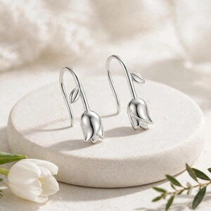 May include: A pair of silver-toned drop earrings shaped like tulip flower buds. Each earring features a curved hook wire, a small leaf detail on the stem, and a smooth, polished metal tulip blossom at the base, reflecting light on its surface.