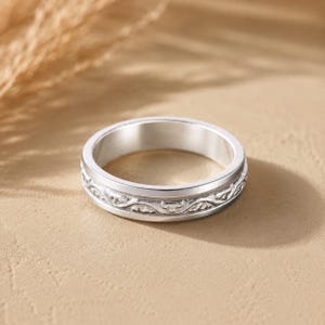 May include: A silver ring with an engraved floral pattern. The ring is circular and has a polished finish. The engraved band features a repeating, symmetrical design. The ring is set against a neutral background.