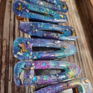 K&ouml;nnte beinhalten: Ein Set mit 10 bunten Haarklammern mit Galaxie-Motiv. Die Klammern bestehen aus klarem Harz mit eingebettetem Glitzer und holografischen Flocken. Jede Klammer zeigt ein anderes himmlisches Design, darunter Sterne, Monde und Planeten.