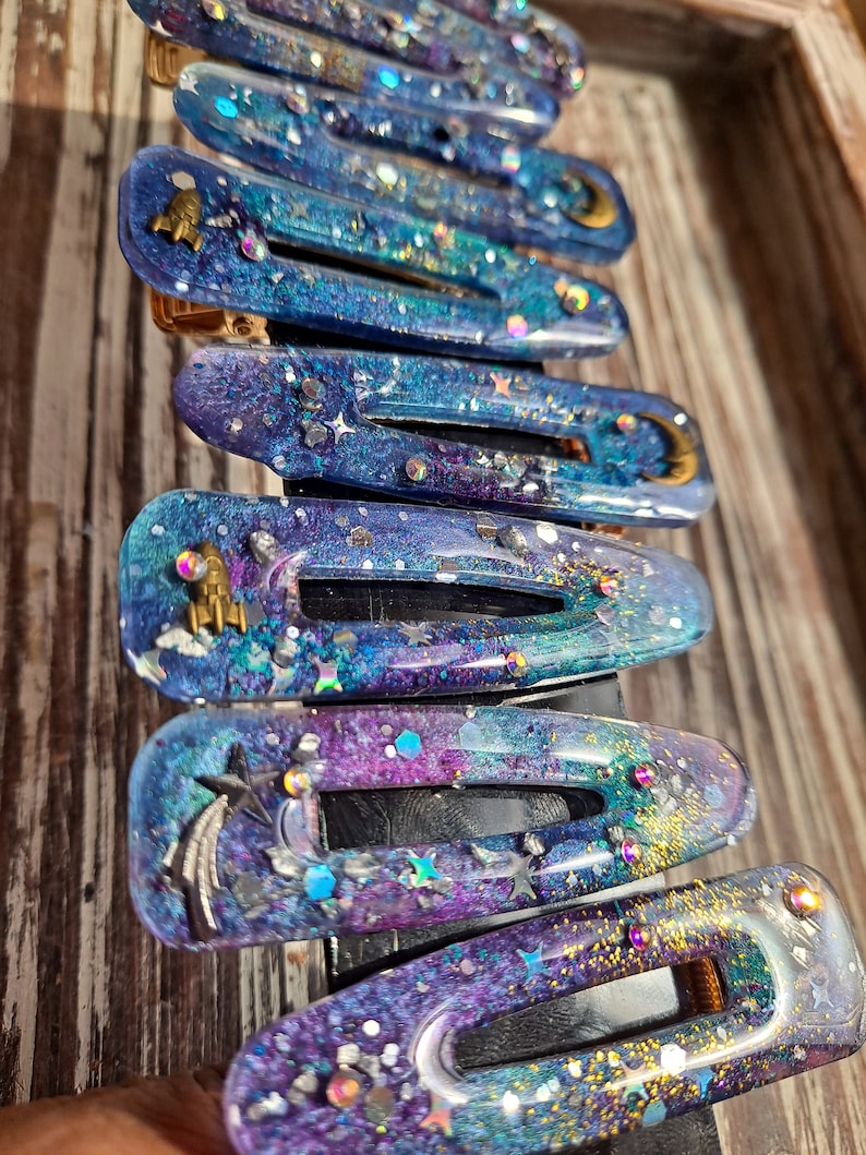 K&ouml;nnte beinhalten: Ein Set mit 10 bunten Haarklammern mit Galaxie-Motiv. Die Klammern bestehen aus klarem Harz mit eingebettetem Glitzer und holografischen Flocken. Jede Klammer zeigt ein anderes himmlisches Design, darunter Sterne, Monde und Planeten.