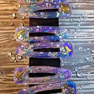 K&ouml;nnte beinhalten: Satz mit sechs lila und blauen Galaxie-Haarklammern mit Glitzer und goldenen Akzenten. Jede Klammer hat einen anderen Weltraum-Anh&auml;nger, darunter eine Rakete, eine Sternschnuppe und einen Planeten.