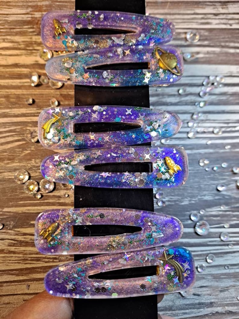 K&ouml;nnte beinhalten: Satz mit sechs lila und blauen Galaxie-Haarklammern mit Glitzer und goldenen Akzenten. Jede Klammer hat einen anderen Weltraum-Anh&auml;nger, darunter eine Rakete, eine Sternschnuppe und einen Planeten.