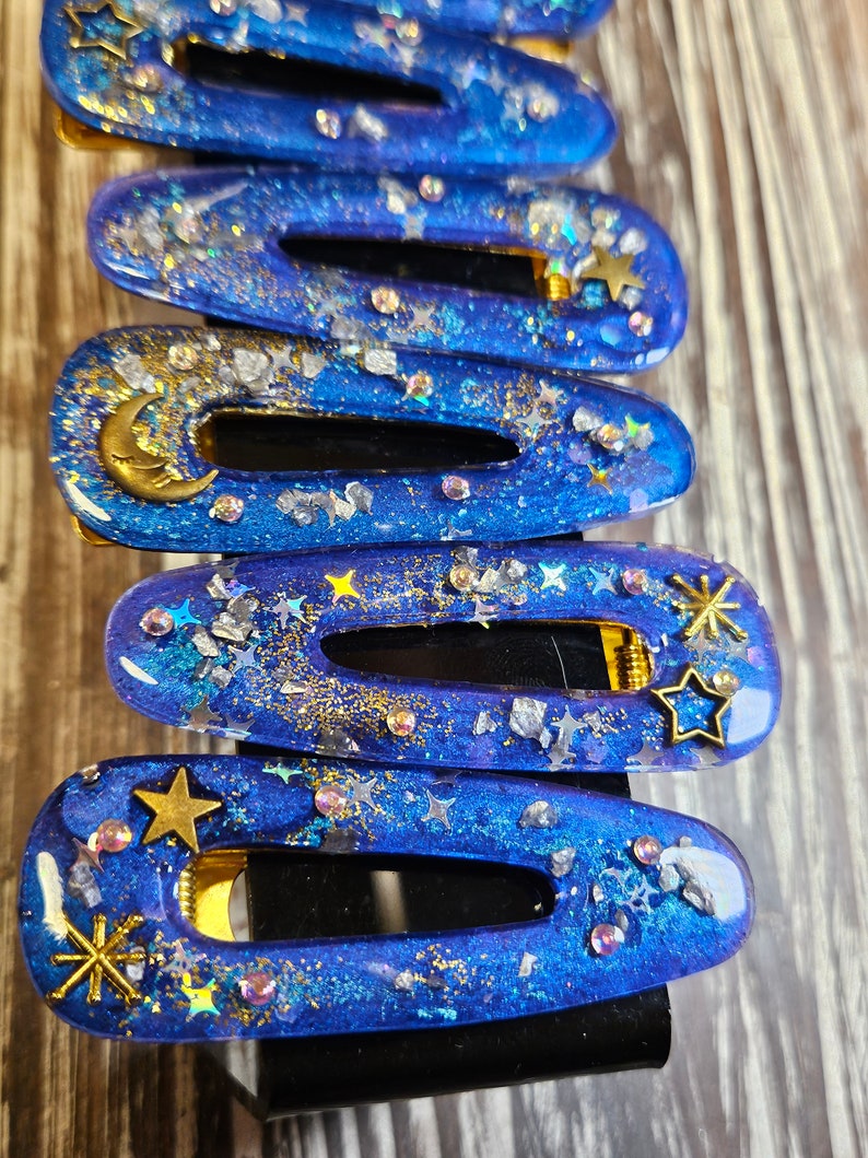 K&ouml;nnte beinhalten: Ein Satz von acht blauen Haarklammern mit goldenem Glitzer und Sternen. Die Klammern sind aus Harz und haben eine klare, gl&auml;nzende Oberfl&auml;che.