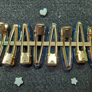 K&ouml;nnte beinhalten: Neun goldene Haarklammern mit einem blauen und schwarzen Galaxie-Design. Die Klammern sind in einer Reihe auf einer grauen Oberfl&auml;che angeordnet.