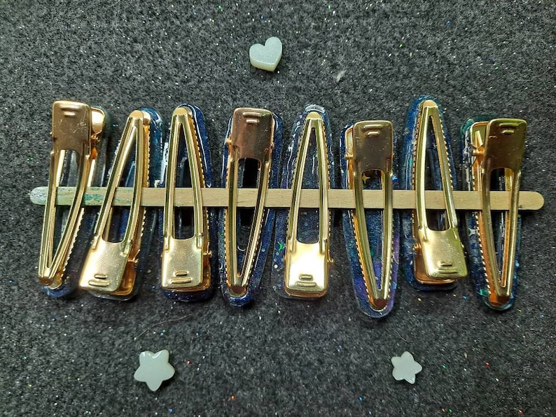 K&ouml;nnte beinhalten: Neun goldene Haarklammern mit einem blauen und schwarzen Galaxie-Design. Die Klammern sind in einer Reihe auf einer grauen Oberfl&auml;che angeordnet.
