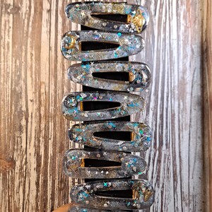 K&ouml;nnte beinhalten: Ein Set mit zehn klaren Haarspangen aus Harz mit goldenen Akzenten und Glitzer. Jede Spange zeigt ein anderes himmlisches Design, darunter Sterne, Monde und Planeten.