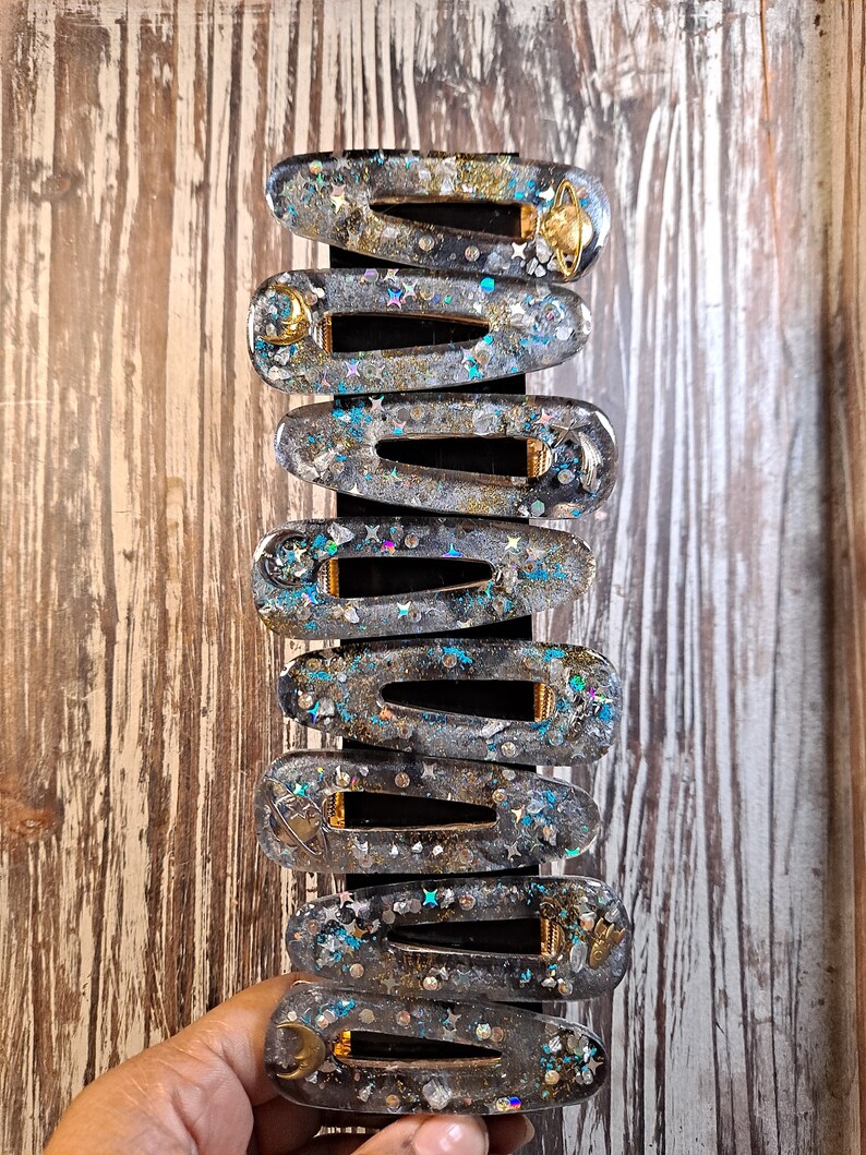 K&ouml;nnte beinhalten: Ein Set mit zehn klaren Haarspangen aus Harz mit goldenen Akzenten und Glitzer. Jede Spange zeigt ein anderes himmlisches Design, darunter Sterne, Monde und Planeten.