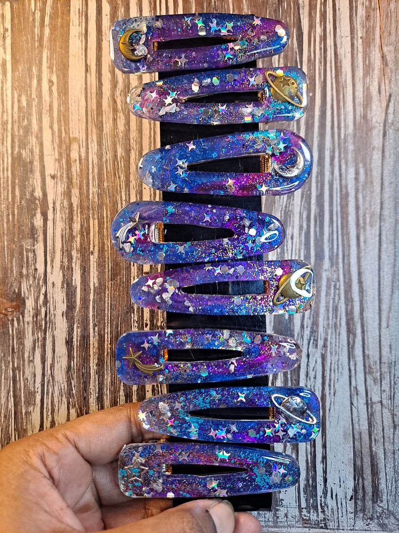 K&ouml;nnte beinhalten: Ein Set mit acht Haarklammern im Galaxie-Design in Lila und Blau mit Glitzer und goldenen Akzenten. Jede Klammer zeigt ein anderes himmlisches Design, darunter Sterne, Monde und Planeten.