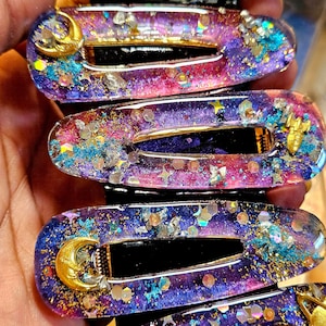 K&ouml;nnte beinhalten: Ein Set mit sechs Haarklammern in Lila, Rosa und Blau mit goldenem Glitzer und einem Halbmond-Anh&auml;nger. Die Klammern sind aus Harz und haben ein galaktisches Design.