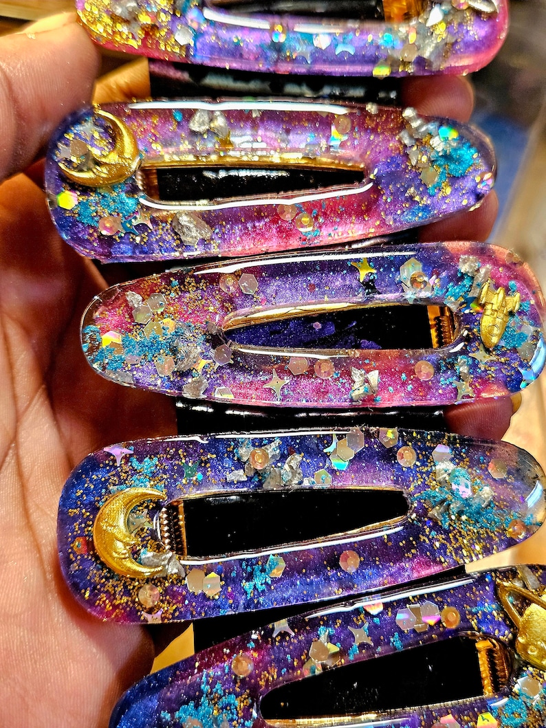 K&ouml;nnte beinhalten: Ein Set mit sechs Haarklammern in Lila, Rosa und Blau mit goldenem Glitzer und einem Halbmond-Anh&auml;nger. Die Klammern sind aus Harz und haben ein galaktisches Design.