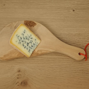 Könnte beinhalten: Ein Stück Blauschimmelkäse auf einem Holzbrett mit einer roten Schnur und einem weißen Etikett.