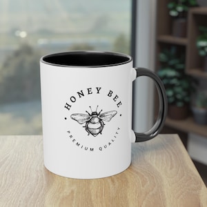May include: White ceramic mug with a black interior and handle. The mug features a detailed illustration of a honey bee, encircled by the words "HONEY BEE PREMIUM QUALITY" in a circular design.