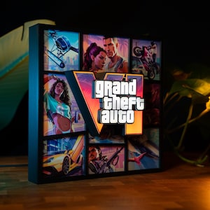 Op de afbeelding: Een vierkante LED-lichtbak met een zwart frame, voorzien van een raster met kleurrijke illustraties uit het videospel Grand Theft Auto VI. Een prominent, verhoogd en verlicht "Grand Theft Auto VI"-logo bevindt zich in het midden en gloeit oranje en wit.