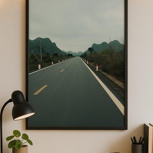 Op de afbeelding: Een ingelijste foto van een lange, rechte weg die door een vallei met groene heuvels en een bewolkte lucht loopt. De weg is geasfalteerd en heeft een witte scheidingslijn. Er staan witte palen langs de zijkanten van de weg.