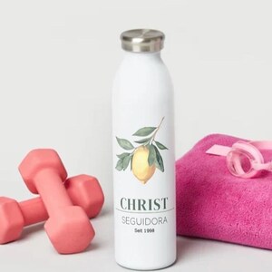 May include: White water bottle with a lemon illustration and the text "CHRIST SEGUIDORA Est. 1998". The bottle is next to pink dumbbells, a pink towel, and pink swimming goggles.
