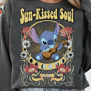 May include: Dark gray sweatshirt featuring a graphic of Stitch playing a ukulele, surrounded by flowers and the text "Sun-Kissed Soul." The design includes a yellow sunburst and the word "Stitch" on a banner.