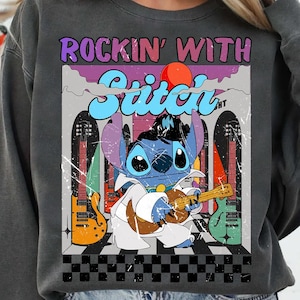 May include: Dark gray sweatshirt featuring a graphic of Stitch playing a guitar, with the text "ROCKIN' WITH Stitch". The design includes guitars and a retro color scheme. The sweatshirt has a crew neck.