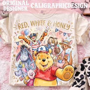 Pode incluir: Uma t-shirt cor de creme com uma impressão gráfica do Winnie the Pooh, Tigre, Ió, Leitão, Coruja e Roo. O texto "RED, WHITE & HONEY" está impresso em dourado sobre as personagens, decoradas com elementos patrióticos e fogo de artifício.