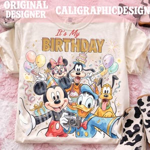 May include: Cream-colored t-shirt featuring a colorful graphic of Mickey Mouse, Minnie Mouse, Goofy, Donald Duck, and Pluto celebrating a birthday. The text "It's My BIRTHDAY" is printed above the characters. The shirt has short sleeves.