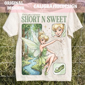 May include: A cream-colored t-shirt featuring a graphic of a blonde fairy with green wings in a forest setting. The text reads "I leave quite an impression" and "SHORT N SWEET". The design includes a close-up of the fairy and a single green shoe.