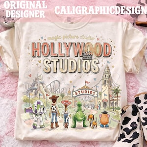 Peut inclure: T-shirt crème avec une illustration colorée de Hollywood Studios, avec le texte "HOLLYWOOD STUDIOS" dans un style vintage. Le design comprend des personnages de dessins animés et une scène de parc à thème. Le t-shirt a également le texte "magic picture studio".