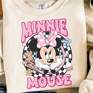 May include: Cream-colored sweatshirt featuring a graphic of Minnie Mouse with a pink bow and the words "MINNIE MOUSE" in pink. The design includes a checkered background, flowers, and a cupcake. The sweatshirt has long sleeves.