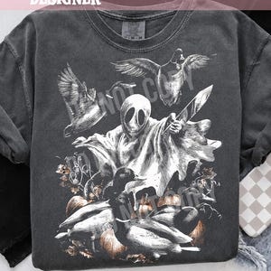 May include: A charcoal gray t-shirt featuring a white graphic print of a ghost holding a knife, surrounded by flying ducks and pumpkins. The design includes a sketch-like illustration style with detailed shading, centered on the front of the garment.