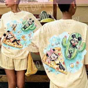 Puede incluir: Una camiseta de color crema con un estampado gráfico de personajes de dibujos animados relajándose sobre una porción de pizza y un flotador de cactus en el agua. El diseño incluye un ratón con sombrero de sol y otro con gafas de sol.