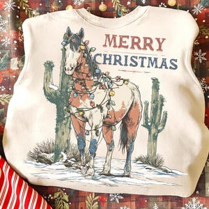 May include: Cream-colored sweatshirt with a graphic of a horse decorated with Christmas lights, standing in a snowy desert scene with cacti. The text "MERRY CHRISTMAS" is above the horse in red and blue.