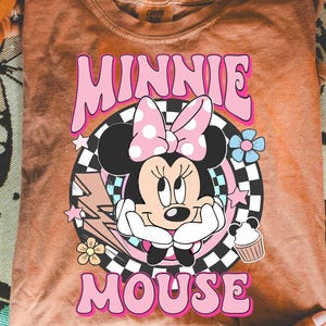 May include: A peach-colored t-shirt featuring a graphic of Minnie Mouse with a pink polka-dot bow, surrounded by flowers, stars, and a lightning bolt. The text "MINNIE MOUSE" is in pink, retro-style lettering.