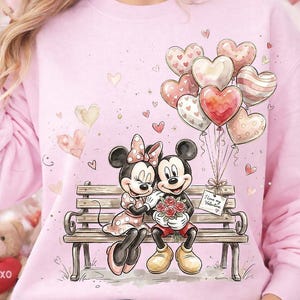 May include: Pink sweatshirt featuring Mickey and Minnie Mouse on a park bench, holding hands and a bouquet of flowers. A cluster of heart-shaped balloons floats above them. The sweatshirt has a Valentine's Day theme.