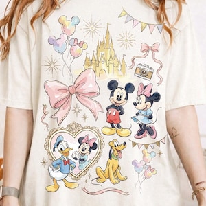 May include: Cream-colored t-shirt featuring a whimsical Disney-themed design. The graphic includes Mickey and Minnie Mouse, Donald Duck, Pluto, a castle, balloons, and bows in pastel colors. A vintage camera and heart-shaped frame are also included.