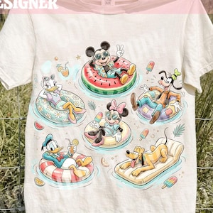 May include: Cream-colored t-shirt featuring a colorful graphic of Disney characters relaxing on inflatable floats in water. Mickey Mouse, Minnie Mouse, Goofy, Donald Duck, Daisy Duck, and Pluto are depicted with drinks and popsicles. Text at the top reads "ORIGINAL CALIGRAPHICDESIGN".