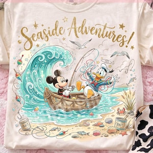 May include: Cream-colored t-shirt featuring a whimsical illustration of Mickey Mouse and Donald Duck fishing in a boat. The design includes a large wave, seagulls, and the text "Seaside Adventures!" in gold glitter. The shirt has a round neckline.