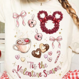 Peut inclure: T-shirt crème avec des illustrations sur le thème de la Saint-Valentin. Les motifs comprennent une couronne en forme de Mickey Mouse, des nœuds roses, des biscuits en forme de cœur et une tasse de chocolat chaud. Le texte "Tis the Valentine Season" est également affiché.