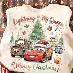 May include: Off-white sweatshirt featuring Lightning McQueen and other Cars characters in a Christmas scene. The design includes a Christmas tree, presents, and the text "Merry Christmas".