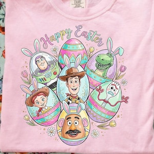 May include: Pale pink t-shirt featuring Toy Story characters in Easter egg designs, each with bunny ears. The words "Happy Easter" are written above the eggs. The shirt is made of a soft material.