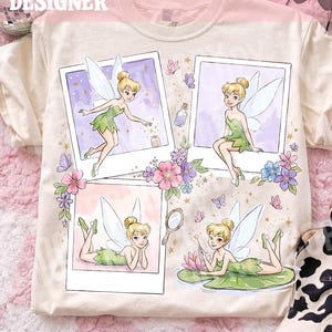 May include: A cream-colored t-shirt featuring four polaroid-style frames with illustrations of a blonde fairy in a green dress. The design includes small butterflies, flowers, a mirror, and a potion bottle, with the text ORIGINAL DESIGNER CALIGRAPHICDESIGN.