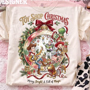 May include: A cream-colored t-shirt featuring a large graphic of a Christmas ornament filled with cartoon characters including a cowboy, a space ranger, a cowgirl, a dinosaur, and a dog. The text reads Toy Shop Christmas and Merry, Bright & Full of Magic.